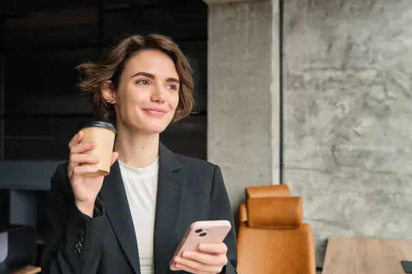 Portrait of woman entrepreneur wearing suit, drinking coffee in her office and holding smartphone, reading message on mobile phone.