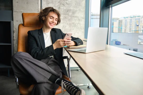 Portrait of businesswoman resting in her office in front of window, using mobile phone, sitting with laptop, looking at smartphone app.