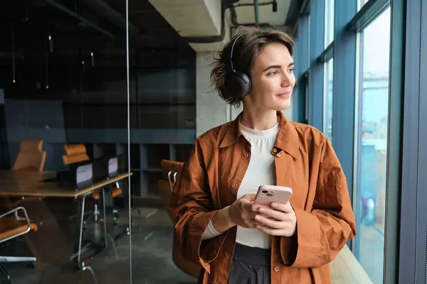 Genç sosyal medya yaratıcısı, kulaklıklı ofis çalışanı, pencerenin yanında akıllı telefonuyla duran ve gülümseyen..