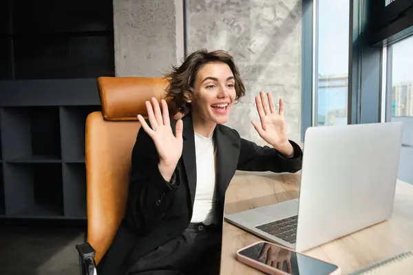 Portrait of happy young businesswoman, waving hand at laptop, joins online meeting or conference, waves hands at computer camera and smiles.