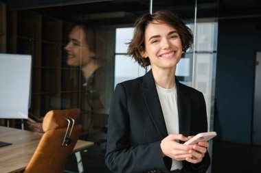 Takım elbiseli bir kadının portresi, ofis odasında akıllı telefonlu ve gülümseyen..