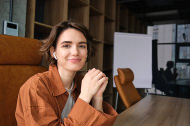 Günlük giysiler içinde genç bir iş kadınının portresi, ofisinde oturan, gülümseyen ve kameraya bakan bir çalışan..