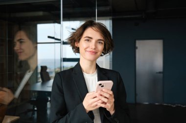 Takım elbiseli bir kadının portresi, ofis odasında akıllı telefonlu ve gülümseyen..