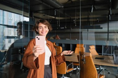 Genç kadın portresi, içerik yaratıcısı, ofisteki Sm yöneticisi, akıllı telefonlu bir video yaratıyor, cep telefonuyla poz veriyor.