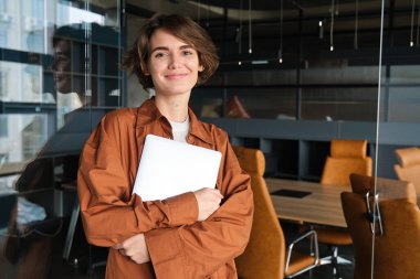 Genç bir kadının portresi, dijital göçebe, ofiste dizüstü bilgisayarla duruyor ve gülümsüyor, işyerinde çalışıyor, çalışma alanında çalışıyor..
