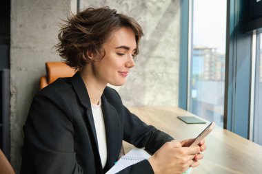 Ofiste çalışan bir kadının portresi, dizüstü bilgisayar ve cep telefonu kullanıyor, birlikte çalışma alanında pencerenin yanında oturuyor..