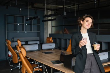 Ofisteki iş kadınının portresi, konferans odasında elinde kahve, cappuccino içiyor ve akıllı telefon kullanıyor..