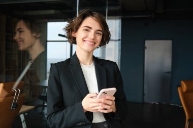Ofiste çalışan başarılı bir iş kadınının portresi, cep telefonuyla cam duvarın yanında duruyor ve gülümsüyor..