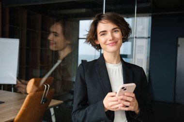 Siyah takım elbiseli şık bir iş kadını, akıllı telefonlu cam duvarın yanında duruyor, ofiste çalışıyor..