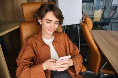 Gündelik giysiler içinde genç bir iş kadınının portresi, akıllı telefonla çalışmak, kablosuz kulaklıkla müzik dinlemek, cep telefonuyla mesajlaşmak, ofiste oturmak..