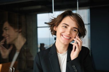 Takım elbiseli bir kadının portresi, ofis odasında akıllı telefonlu ve gülümseyen..