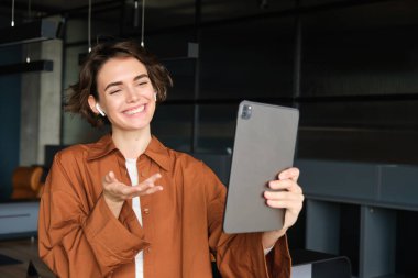 İş ortağıyla konuşan bir kadının portresi dijital tabletle çevrimiçi görüşmeye bağlanıyor, ofiste dikiliyor, günlük kıyafetler giyiyor..