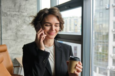 Genç bir girişimcinin resmi bir telefona cevap veriyor, ofis penceresinin yanında kahve içiyor ve konuşurken gülümsüyor..