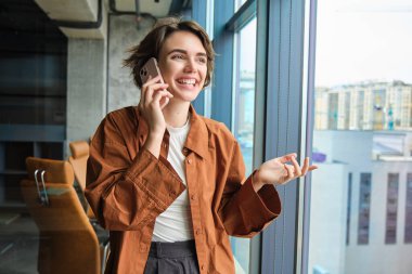 Genç satış elemanı portresi, ofis çalışanı, müşteri, iş kadını cep telefonuyla konuşuyor ve iş yerinin penceresinin yanında duruyor..