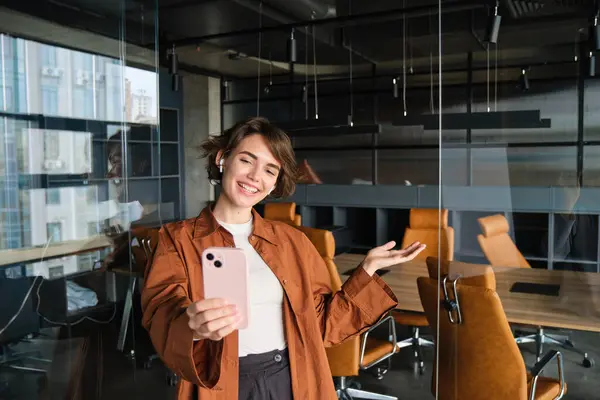 Image of woman, start up manager, creates content for online social media of company, showing office to someone, pointing at conference room, taking selfie with smartphone.