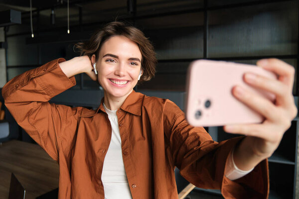 Portrait of young woman, social media content creator in office, posing for selfie, taking pictures on smartphone, posing for photo on mobile phone.