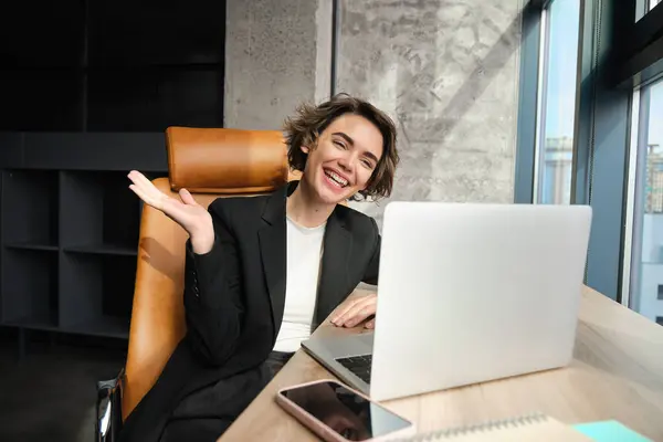 Portrait of corporate woman, ceo manager joins online meeting or webinar, saying hello at laptop camera, waving hand to greet coworkers, sitting in office.