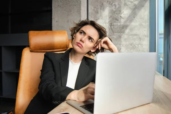 Portrait of thinking woman, corporate businesswoman in her office, sitting with laptop, looking up with thoughtful face, pondering.
