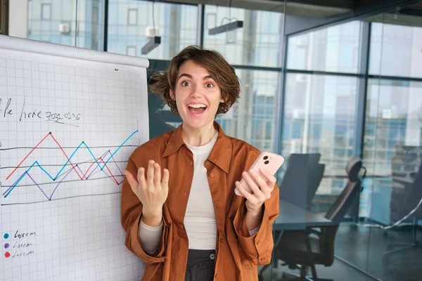 Portrait of woman expresses excitement, office worker dances near diagram with company stats, holds smartphone, reacts to wonderful news or achievement.