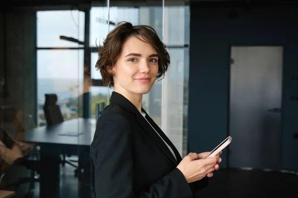 Image of corporate woman, saleswoman in business company, standing in black suit, holding smartphone, calling client.