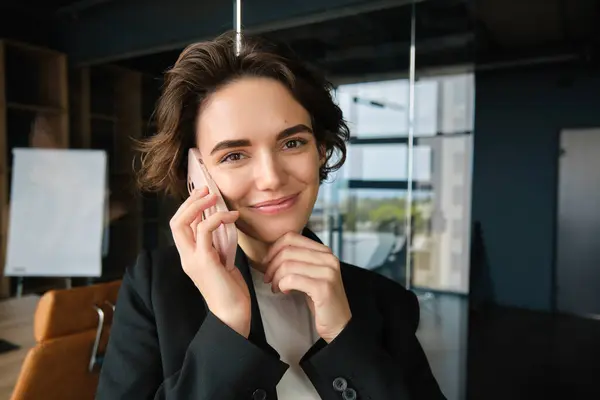 Portrait of young corporate woman, businesswoman in suit, answers phone call and smiles, stands in office conference room. Business and people concept