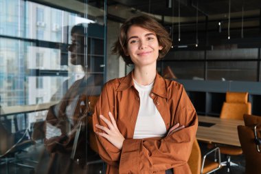 Kendine güvenen genç iş kadını portresi, ofisinde poz veren dijital göçebe, günlük kıyafetler giymiş ve gülümsüyor..