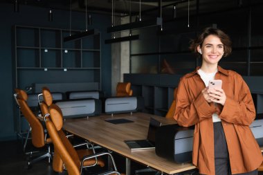 Günlük kıyafetlerle gülümseyen, akıllı telefonuyla ofiste duran, kablosuz kulaklıkla cep telefonuyla konuşan genç bir kadın..