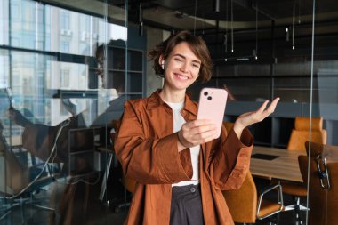 Genç bayan girişimcinin görüntüsü, dijital göçebe ofiste duruyor, kablosuz kulaklıklarla çevrimiçi toplantılara bağlanıyor, video sohbetleri, akıllı telefon kullanıyor, ortak çalışma alanında poz veriyor.