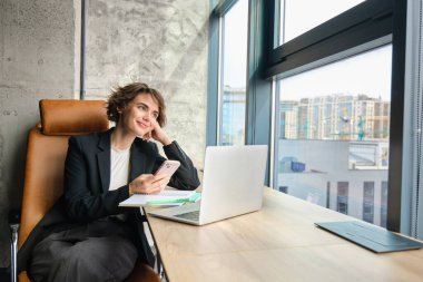 Takım elbiseli genç satış elemanının resmi, dizüstü bilgisayarla çalışıyor, ofisinde akıllı telefonuyla oturuyor, mesajlara cevap veriyor, cep telefonundan kitap okuyor, uygulamayı kullanıyor..