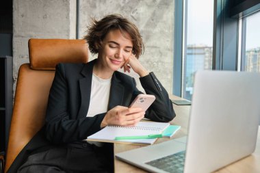 Ofisinde çalışan bir kadının portresi. Başarılı iş kadını dizüstü bilgisayarı ve telefonuyla oturuyor, gülümsüyor ve mutlu görünüyor..