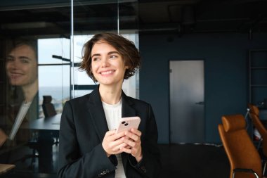 Siyah takım elbiseli şık bir iş kadını, akıllı telefonlu cam duvarın yanında duruyor, ofiste çalışıyor..