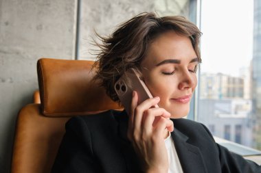 Gülümseyen esmer kadın, cep telefonuyla konuşan iş kadını, telefonla konuşan ve gülümseyen deri koltukta oturan,.