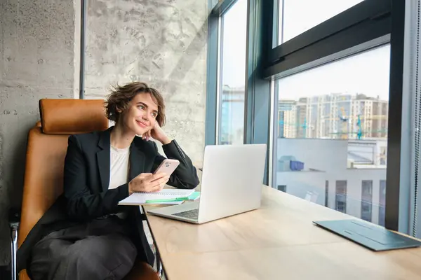 Takım elbiseli genç satış elemanının resmi, dizüstü bilgisayarla çalışıyor, ofisinde akıllı telefonuyla oturuyor, mesajlara cevap veriyor, cep telefonundan kitap okuyor, uygulamayı kullanıyor..