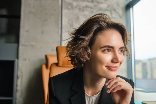 Portrait of corporate woman looking outside window in her office, smiling with confidence. Business lifestyle