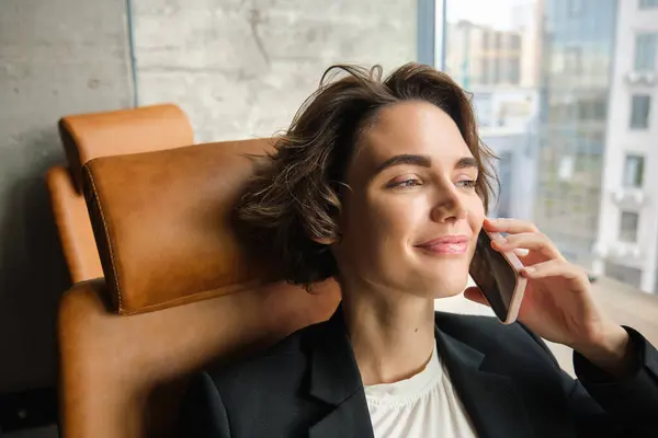 Portrait of business woman in suit, sitting in her office and answering a phone call with pleased smile, having a negotiation over the telephone conversation.