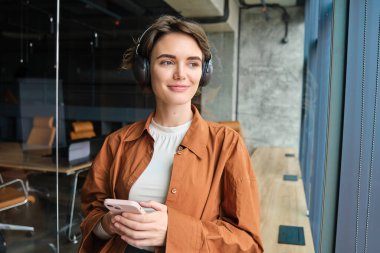 Dijital göçebe portresi, kablosuz kulaklıklı kadın yönetici, pencerenin yanında duruyor, cep telefonu tutuyor..