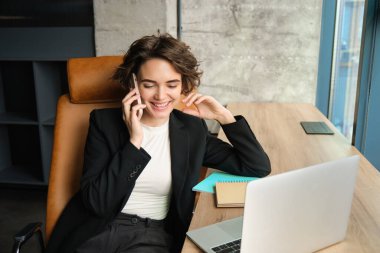 Ofisinde oturan, telefonlara cevap veren, akıllı telefonunu ve laptopunu kullanan, pencerenin yanında çalışan genç bir iş kadınının portresi..