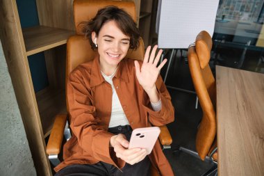 Günlük giysiler içinde bir kadın, yönetici ofisinde oturuyor ve çalışıyor, online toplantısı var, akıllı telefon uygulamasından görüntülü sohbete bağlanıyor..