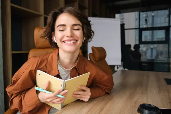 Portrait of happy smiling female entrepreneur, employee in office, sitting at the table, holding planner, making notes, writing information during a meeting and looking happy.
