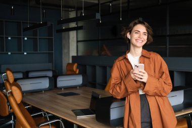 Genç modern kadın, ofis çalışanı konferans odasında akıllı telefon ve kablosuz kulaklıkla duruyor..