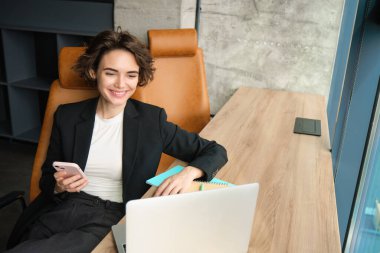 Satış elemanı telefonuna cevap veriyor, akıllı telefondan müşteriyle konuşuyor, bilgisayarlı bir ofiste çalışıyor, pencerenin yanında oturuyor..