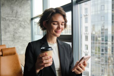 Kadın girişimci portresi, iş yerinde dikilip elinde akıllı telefon, pencerenin yanında kahve içiyor..