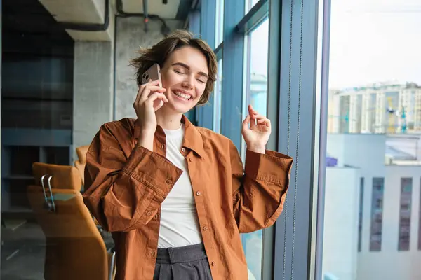 Portrait of working woman, social media manager in office, talking on mobile phone, having conversation on telephone, standing next to the window.