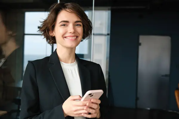 Portrait of saleswoman working in company, standing in office near glass wall, holding mobile phone and smiling, calling client, arranging a business meeting.