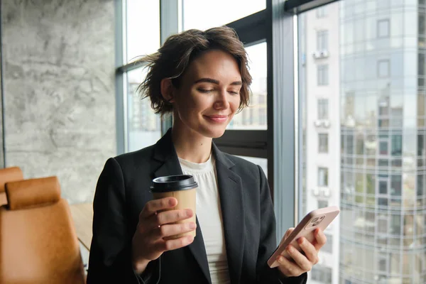 Portrait of female entrepreneur, standing in business office and holding smartphone, drinking coffee next to the window.