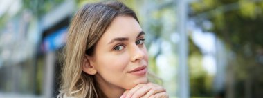 Genç bir kadının yakın portresi, saçlarını kulaklarının arkasına sıkıştırmak, cilveli görünmek, gülümsemek, açık havada mavi gömlekle oturmak..