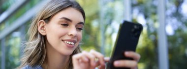 Gülümseyen kız dışarıda oturuyor, akıllı telefona bakıyor, cep telefonundan sipariş veriyor, uygulamayı kullanıyor..