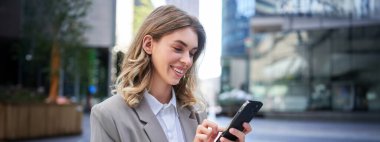 Gülümseyen genç iş kadını dışarıda dururken cep telefonu kullanıyor. Kurumsal insan kavramı.