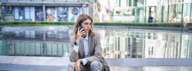 Takım elbiseli şık bir iş kadını, ofis binalarının dışındaki çeşmenin yanında oturuyor, cep telefonuyla konuşuyor. Şirket kadını dışarıda birini bekliyor..