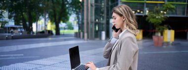 Cep telefonuyla konuşan, dizüstü bilgisayarla çalışan, şehir merkezinde oturan, takım elbiseli bir kadın..